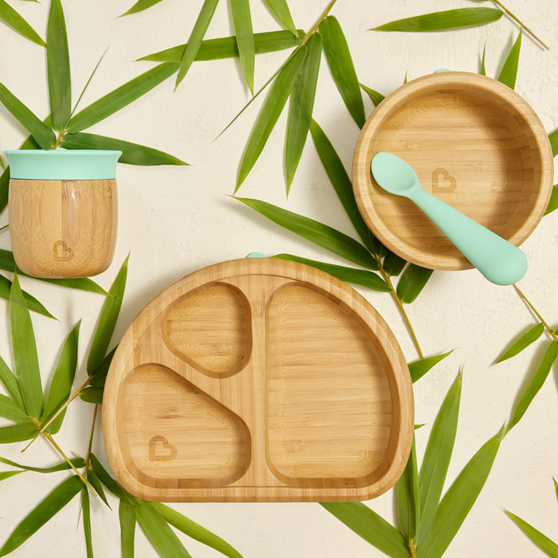 A collection of bamboo weaning products on a bed of bamboo leaves, including a bamboo open cup, bamboo suction bowl and spoon, and bamboo suction divider plate