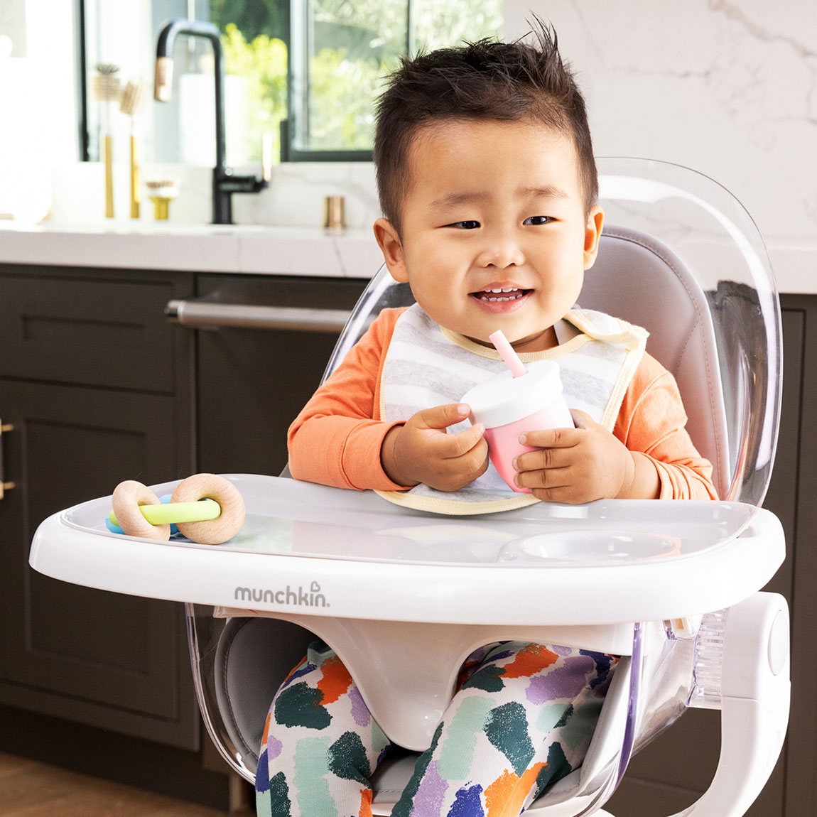 A smiling toddler sitting in a Munchkin High Chair holding a C'est Silicone sippy straw cup